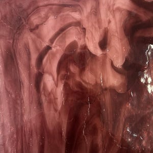 May include: Close-up of a sheet of stained glass with a swirling pattern in shades of mauve and burgundy. The glass has a translucent quality, with light passing through the varying tones, creating a dynamic visual effect.