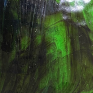 May include: Close-up of a sheet of stained glass with a swirling pattern. The glass features shades of green and black, creating a textured, abstract design. The colors blend together, with areas of deep, dark green and lighter, brighter green.