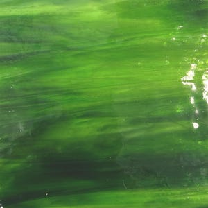 May include: A close-up of a sheet of green stained glass. The glass has a textured, flowing pattern with varying shades of green, from light to dark. The light reflects off the surface, creating highlights and shadows.