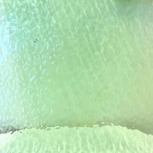 May include: An aerial view of shallow, clear water with a sandy bottom. The water has a light green hue, with subtle wave patterns and ripples visible on the surface. The sandy bottom is visible near the edge.