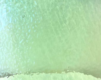 May include: An overhead shot of shallow, clear water with a sandy base. The water has a pale green tint, with gentle wave patterns and ripples on the surface. The sandy base is visible near the edge.