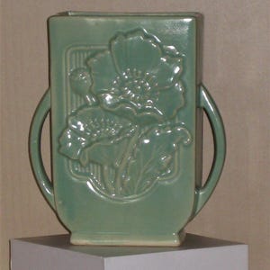 May include: A sage green ceramic vase with a rectangular body and two curved side handles. The front features a raised relief design of two large poppy flowers with leaves and a bud, set against a vertical striped rectangular background.