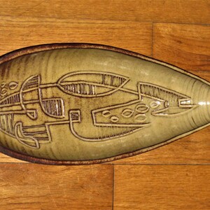 May include: An elongated, leaf-shaped ceramic dish featuring a muted olive-green glaze with a dark brown rim. The surface is decorated with incised, abstract geometric line patterns and circular motifs that reveal the darker clay body underneath.