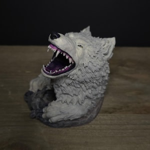 May include: A gray wolf figurine with an open mouth, revealing sharp white teeth and a purple interior. The wolf is in a seated position, with detailed fur texture. The figurine is on a wooden surface.