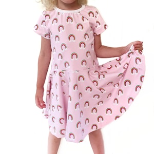 Puede incluir: Un vestido rosa con estampado de arcoíris. El vestido tiene mangas cortas y un escote redondo.