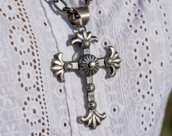 Könnte beinhalten: Ein silberfarbener Kreuzanhänger mit aufwendigen Details hängt an einer klobigen Kette. Das Kreuz hat ein zentrales kreisförmiges Design und dekorative Verzierungen. Die Halskette liegt auf weißem Lochmusterstoff.