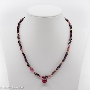 Op de afbeelding: Een ketting gemaakt met kleine, donkerrode en roze kralen met een grotere, druppelvormige roze edelsteen hanger.