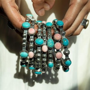 Puede incluir: Una colección de brazaletes plateados, cada uno adornado con piedras preciosas turquesas y rosas. Los brazaletes tienen un diseño detallado y texturizado con detalles de estrellas y cuentas. Las piedras preciosas son de forma ovalada y están engastadas en metal pulido.