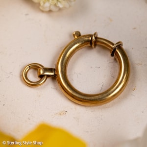 Puede incluir: Un cierre de resorte de metal en tono dorado que presenta un aro principal circular con un mecanismo de resorte y una anilla más pequeña adjunta. El componente tiene un acabado liso y pulido, diseñado para proyectos de joyería.