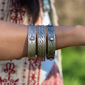 May include: Three silver-toned metal cuff bracelets with detailed engraved patterns. Each bracelet has a unique design, including geometric and floral patterns. The bracelets are displayed on an arm, highlighting their width and detail. The bracelets are approximately 2.5 cm wide.