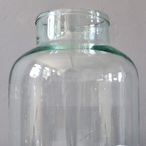 May include: A large, clear glass jar with a wide mouth and a slightly green tint. The jar is empty and sitting on a white fabric surface.