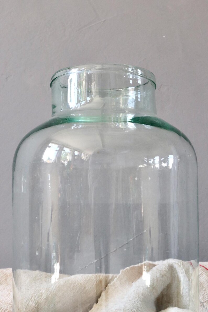 May include: A large, clear glass jar with a wide mouth and a slightly green tint. The jar is empty and sitting on a white fabric surface.
