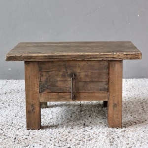 May include: A rustic, square wooden side table with a weathered appearance. The table features a single drawer with a metal handle and is supported by four sturdy legs. The wood has a natural, aged finish, showcasing the grain and knots.