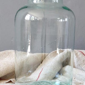 May include: A large, clear glass jar with a slightly green tint. The jar has a wide mouth and a rounded bottom. It is sitting on a white fabric surface with red stripes.