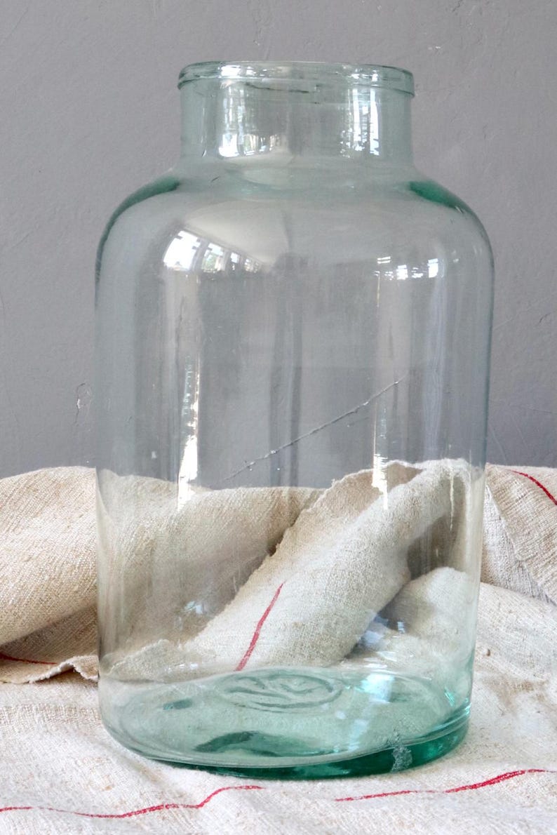 May include: A large, clear glass jar with a slightly green tint. The jar has a wide mouth and a rounded bottom. It is sitting on a white fabric surface with red stripes.