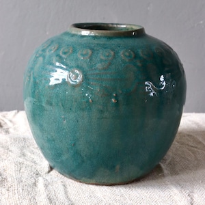 Könnte beinhalten: Eine türkisfarbene, runde Keramikvase mit leicht strukturierter Oberfläche. Die Vase weist eine Reihe von geprägten Wirbelmustern um den oberen Teil auf. Die Öffnung ist in einem dunkleren Grünton gehalten.