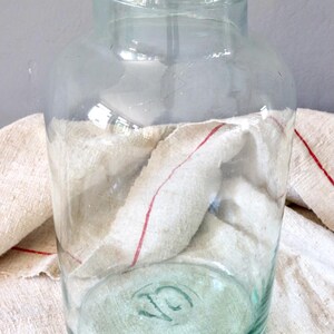 May include: A large, clear glass jar with a green tint. The jar has a wide mouth and a rounded bottom. The jar is sitting on a white and red striped cloth.