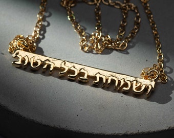 May include: A gold necklace with a bar pendant inscribed with the Hebrew words "Shehecheyanu" in a modern font. The necklace is resting on a grey concrete heart-shaped surface with dried rose petals scattered around it.