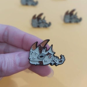 May include: A close-up of a white and brown enamel pin depicting a cartoon creature with a spiky back and horns. The creature has a single large eye and a small mouth. The pin is being held in a hand.
