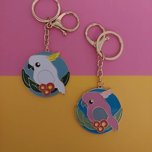 May include: Two gold keychains with round enamel charms featuring cartoon cockatoos. One charm is blue with a white cockatoo and the other is blue with a pink cockatoo. Both charms have green leaves and red flowers.