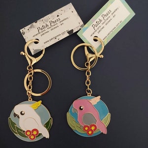 May include: Two gold keychains with enamel charms featuring a white cockatoo and a pink cockatoo on a blue background with green leaves and red flowers. The keychains are from Patch Press, a company that sells enamel pins, keychains, jewelry, patches, stickers, and art.