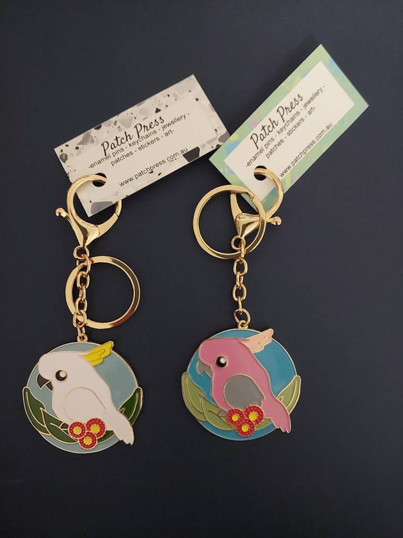 May include: Two gold keychains with enamel charms featuring a white cockatoo and a pink cockatoo on a blue background with green leaves and red flowers. The keychains are from Patch Press, a company that sells enamel pins, keychains, jewelry, patches, stickers, and art.