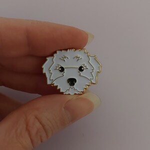 May include: A white enamel pin with a gold outline depicting a white dog with black eyes and nose. The dog is looking directly at the viewer.