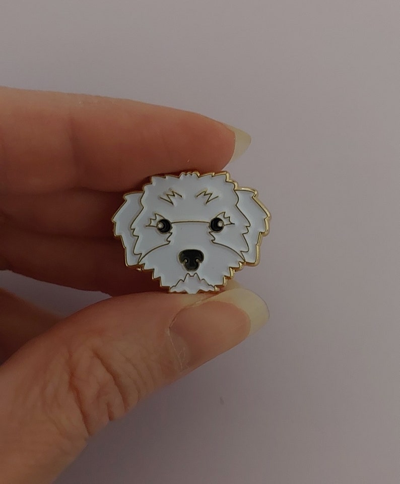 May include: A white enamel pin with a gold outline depicting a white dog with black eyes and nose. The dog is looking directly at the viewer.