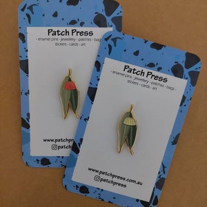 Könnte beinhalten: Zwei Emaille-Pins mit einer stilisierten Illustration einer Gummibaum-Blume. Die Pins befinden sich auf blau-schwarz gemusterten Karten mit dem Text "Patch Press - enamel pins - jewellery - patches - bags - stickers - cards - art" und Website-Informationen.