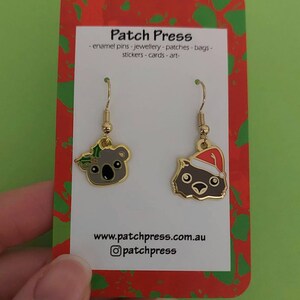 May include: A pair of gold-tone earrings featuring a koala wearing a holly wreath and a wombat wearing a Santa hat. The earrings are displayed on a red and green card with the text "Patch Press - enamel pins - jewellery - patches - bags - stickers - cards - art - www.patchpress.com.au @patchpress".