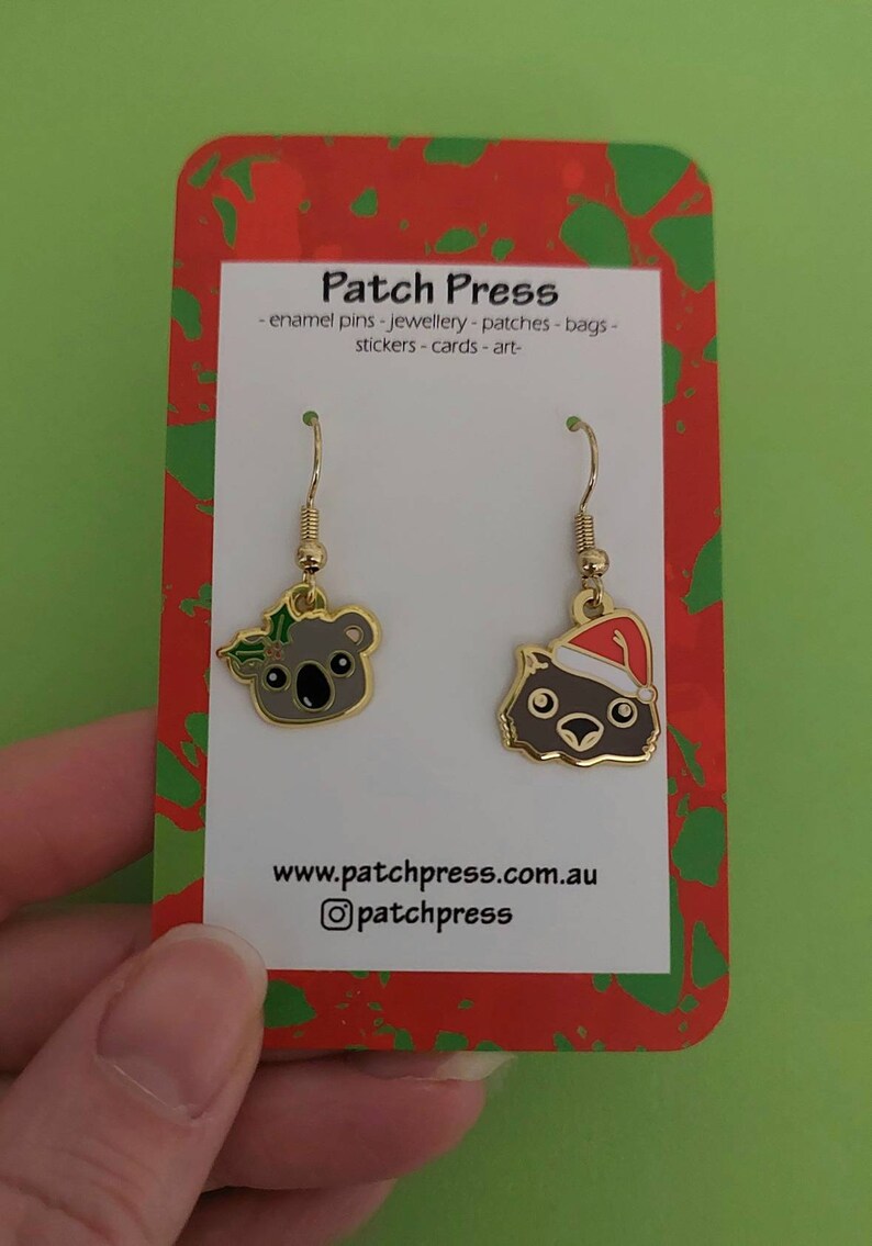 May include: A pair of gold-tone earrings featuring a koala wearing a holly wreath and a wombat wearing a Santa hat. The earrings are displayed on a red and green card with the text "Patch Press - enamel pins - jewellery - patches - bags - stickers - cards - art - www.patchpress.com.au @patchpress".