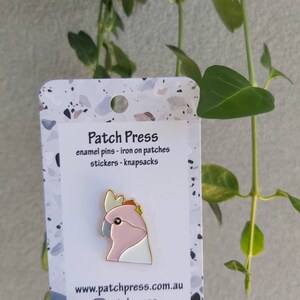 May include: A pink and white enamel pin of a cockatoo bird with a gold outline. The pin is on a white card with the text "Patch Press" and "enamel pins - iron on patches stickers - knapsacks" in black text. The card also has the website "www.patchpress.com.au" and the Instagram handle "@patchpress" in black text.