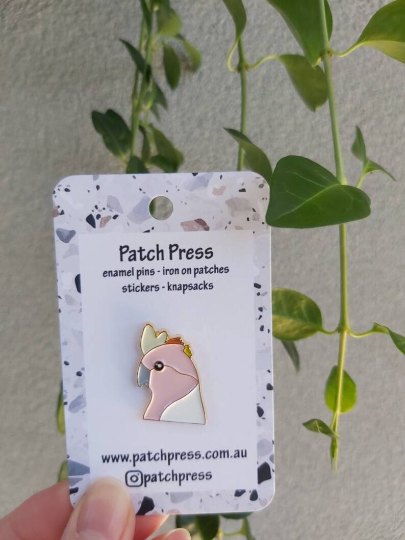 May include: A pink and white enamel pin of a cockatoo bird with a gold outline. The pin is on a white card with the text "Patch Press" and "enamel pins - iron on patches stickers - knapsacks" in black text. The card also has the website "www.patchpress.com.au" and the Instagram handle "@patchpress" in black text.