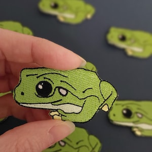 May include: Five green embroidered frog patches with black eyes. The frog patches are in various poses, including one with a winking eye.