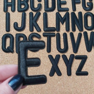 May include: Black felt letters of the alphabet, A through Z, are arranged on a corkboard. The letter E is being held by a hand.