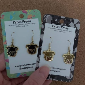 Puede incluir: Dos pares de pendientes de tono dorado con un dise&ntilde;o de pug de dibujos animados. Los pendientes se muestran en una tarjeta blanca con el texto "Patch Press" e informaci&oacute;n del sitio web.
