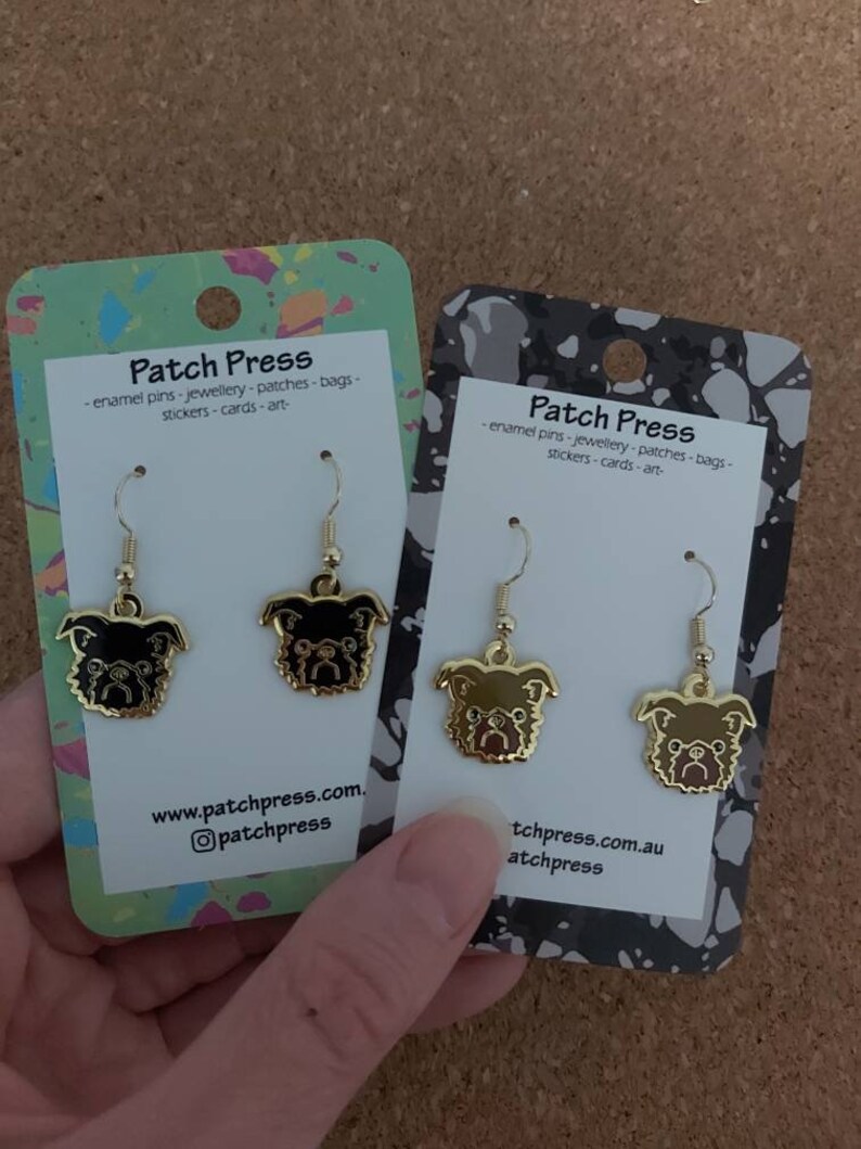 Puede incluir: Dos pares de pendientes de tono dorado con un dise&ntilde;o de pug de dibujos animados. Los pendientes se muestran en una tarjeta blanca con el texto "Patch Press" e informaci&oacute;n del sitio web.