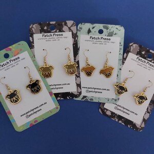 Puede incluir: Cuatro pares de pendientes de tono dorado con un dise&ntilde;o de carlino. Los carlinos est&aacute;n representados en negro, marr&oacute;n y blanco. Los pendientes se muestran en tarjetas con el texto "Patch Press" y una direcci&oacute;n de sitio web.