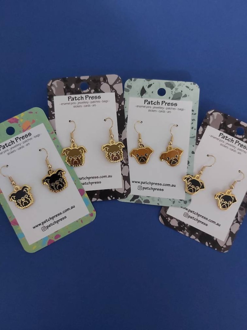 Puede incluir: Cuatro pares de pendientes de tono dorado con un dise&ntilde;o de carlino. Los carlinos est&aacute;n representados en negro, marr&oacute;n y blanco. Los pendientes se muestran en tarjetas con el texto "Patch Press" y una direcci&oacute;n de sitio web.