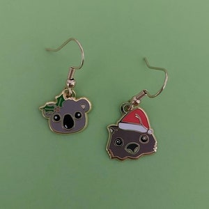 May include: A pair of gold dangle earrings featuring a koala wearing a holly wreath and a wombat wearing a Santa hat. The earrings are on a green background.