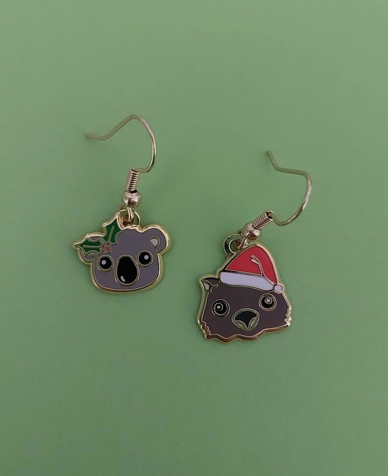 May include: A pair of gold dangle earrings featuring a koala wearing a holly wreath and a wombat wearing a Santa hat. The earrings are on a green background.