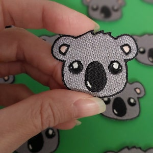 May include: A grey embroidered koala bear patch with black eyes and a black nose. The patch is held in a hand against a green background.