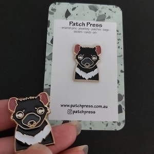 May include: A black and white enamel pin of a Tasmanian devil with a gold outline. The pin is on a white card with the text "Patch Press" and the website address "www.patchpress.com.au".