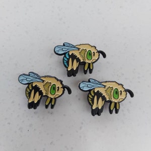 K&ouml;nnte beinhalten: Drei Emaille-Pins mit Cartoon-Bienen mit schwarzen K&ouml;rpern, gelben und braunen Streifen, gr&uuml;nen Augen und blauen Fl&uuml;geln.