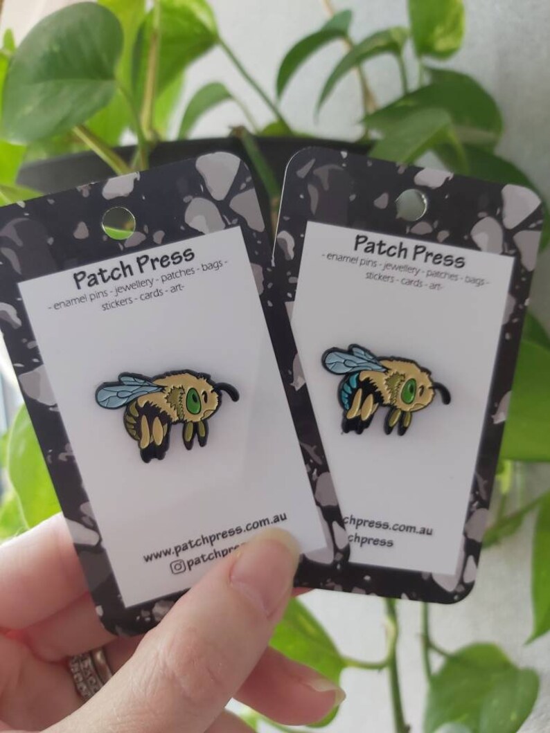 K&ouml;nnte beinhalten: Zwei Emaille-Pins mit einer Cartoon-Biene mit blauen Fl&uuml;geln, gelbem K&ouml;rper und schwarzen Streifen. Die Pins befinden sich auf wei&szlig;en Karten mit schwarzen R&auml;ndern und dem Text "Patch Press" und "www.patchpress.com.au".
