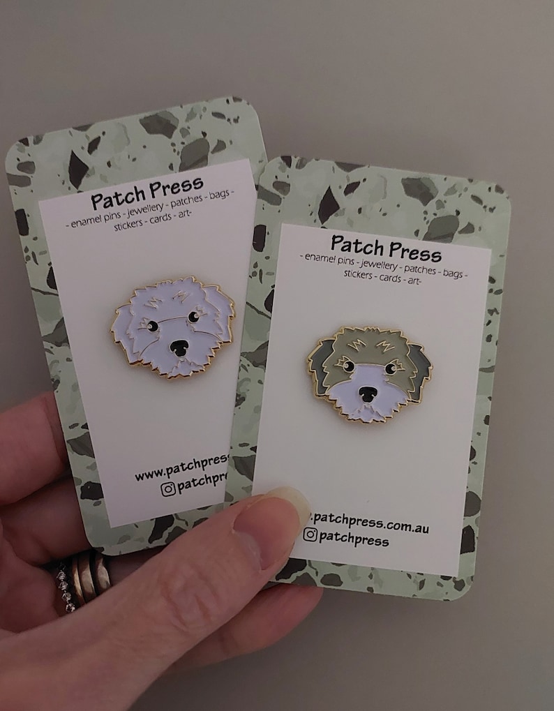 May include: Two white cards with green and brown speckled backgrounds. Each card features a white enamel pin of a dog with a white and gray fur pattern. The text on the cards reads "Patch Press - enamel pins - jewellery - patches - bags - stickers - cards - art - www.patchpress.com.au @patchpress".