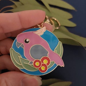 May include: A pink and grey cockatoo bird keychain with a blue background and gold accents. The bird is surrounded by green leaves and red flowers.