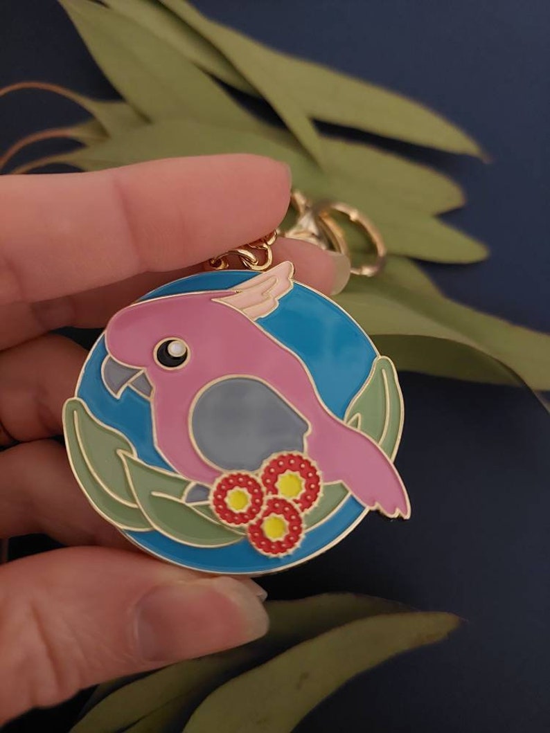 May include: A pink and grey cockatoo bird keychain with a blue background and gold accents. The bird is surrounded by green leaves and red flowers.