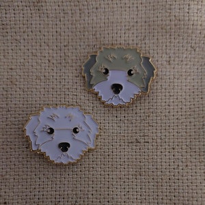 May include: Two enamel pins featuring cartoon dog faces. The dog on the left has white fur and the dog on the right has gray fur.
