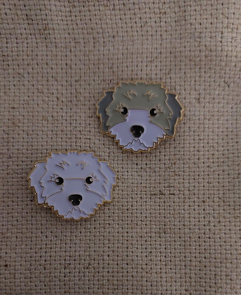 May include: Two enamel pins featuring cartoon dog faces. The dog on the left has white fur and the dog on the right has gray fur.